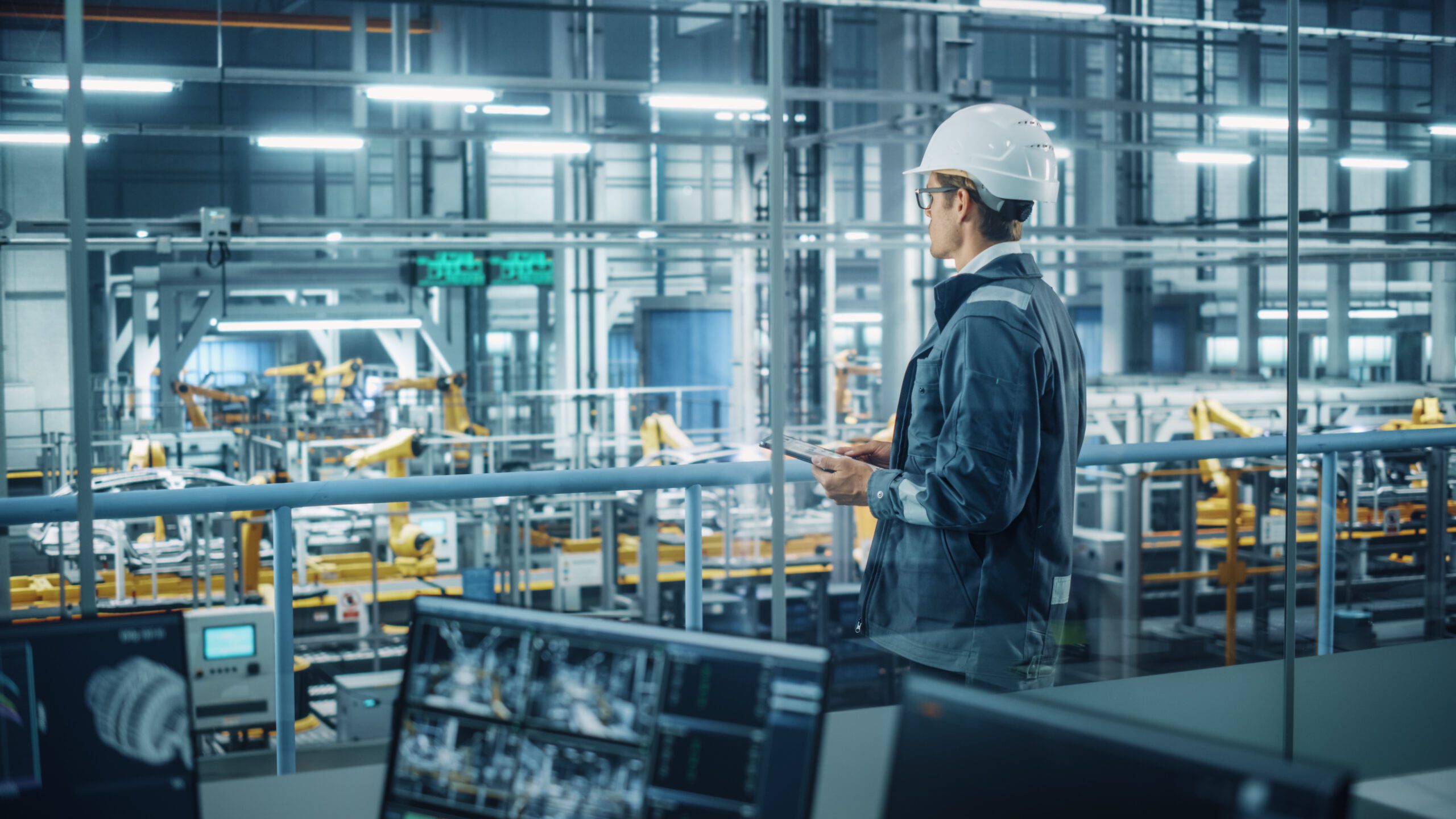 Handsome Engineer in Uniform and Hard Hat Using Tablet Computer at a Car Assembly Plant. Industrial Specialist Working on Vehicle Design, Overlooking Production in Technological Facility.